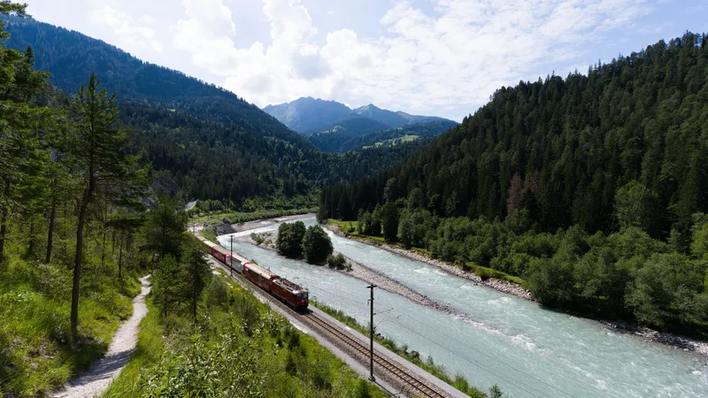 A red Rhaetian Railway train follows a turquoise river through a forested Swiss alpine valley