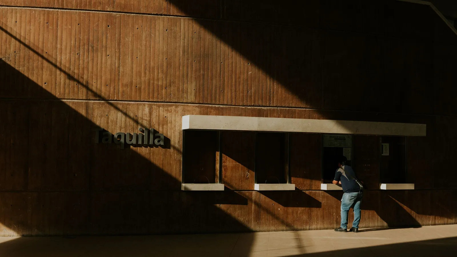Een reiziger bij een stationsloket met het opschrift Taquilla, terwijl de zon lange schaduwen op de muur werpt