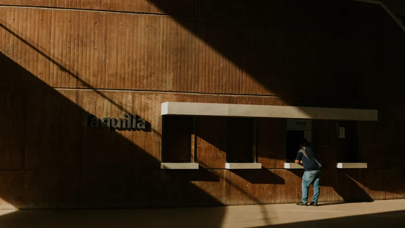 A traveller at a station ticket booth marked Taquilla, sunlight casting long shadows on the wall